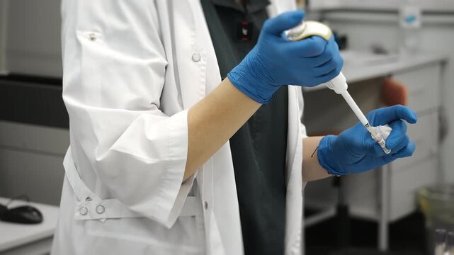 Close up of laboratory technician in white coat and blue gloves using micropipette for blood sample analysis in modern medical laboratory indoors.