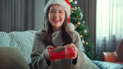 Asian woman wearing Santa hat smiling and holding Christmas gift box with decorated tree in background, joyful holiday celebration and festive season concept, Christmas greeting and happiness at home.