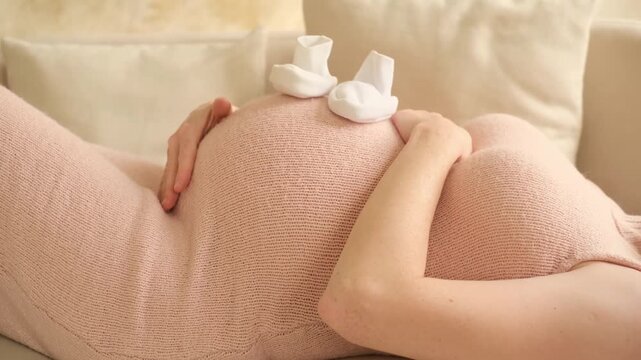 a pregnant woman pats her stomach while holding baby booties or socks, a woman waiting for a baby to be born, close-up of a pregnant woman's stomach