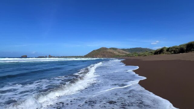 The sea in Jember, East Java. Indonesia. Watu Ulo beach with dark sand, mountains and coral rocks towering above the beach. The raging blue Indian ocean on a sunny day. 4К