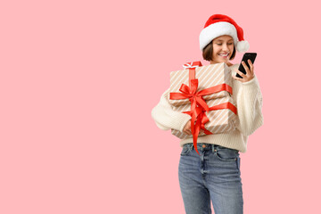Young happy woman in Santa hat with gift boxes and mobile phone on pink background. Christmas celebration