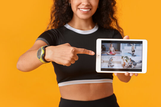 Sporty young African-American female coach pointing at tablet computer on yellow background. Concept of online training