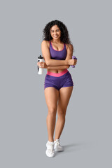 Fototapeta premium Sporty young African-American female coach with water bottle and dumbbells on white background