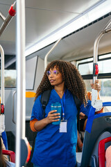 African nurse in scrubs standing on a city bus with reusable cup and id badge, commuting to work, focused and ready