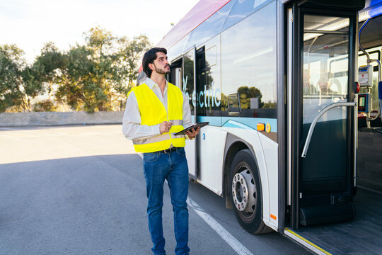 Quality inspector checking a modern public electric bus in an open air depot, holding a tablet and wearing a safety vest - Powered by Adobe