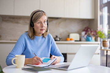 Senior woman engaging in remote work or e-learning from her home kitchen, wearing a headset, taking notes in a notebook while looking at her laptop screen