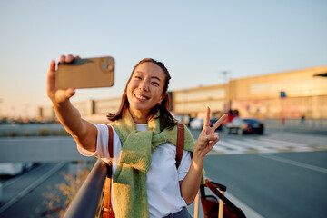 Happy woman smiling and making a peace sign, taking a selfie with a phone at an airport, anticipating travel or vacation