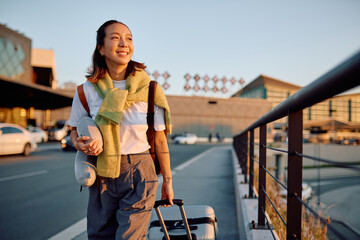 Happy young asian woman traveler walking with suitcase and neck pillow at an airport at sunset, beginning new journey