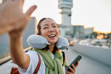 Smiling Asian woman traveling, wearing a neck pillow and scarf, doing a high five, ready to go