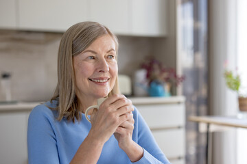 Fototapeta premium Senior woman smiling warmly while holding a warm mug with both hands, enjoying a quiet moment of relaxation and contemplation in her bright home kitchen