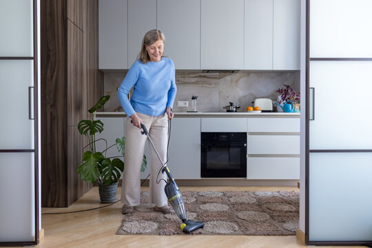 Happy senior woman cleaning a rug with a modern stick vacuum cleaner in her contemporary kitchen, performing daily chores and maintaining a tidy living space