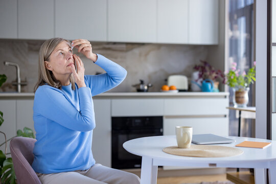 Senior grey-haired woman carefully applying prescribed eye drops into her eye for vision care, dry eye relief, and routine self-health treatment at home