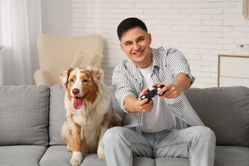 Young man playing video games with cute Australian Shepherd dog and sitting on sofa in living room