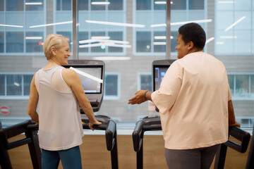 Obraz premium Caucasian senior woman and Black middle aged woman using treadmills in modern gym, both standing side by side and engaging in conversation while exercising indoors near large windows