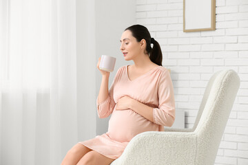 Beautiful young pregnant woman with cup of tea sitting in armchair in living room