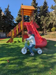 Toddler on a balance bike riding on a sunny autumn day at a colorful playground.

