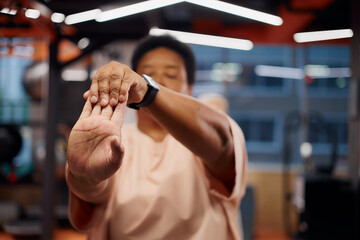 Middle aged Black woman stretching arm forward in gym, focusing on hand and fingers, blurred face in background, wearing smartwatch, engaging in fitness activity indoors