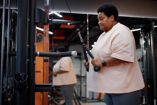 Middle aged Black woman exercising on cable machine in gym, gripping rope attachment and performing triceps pushdown, focused on strength training, wearing smartwatch - Powered by Adobe