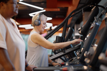 Fototapeta premium Senior Caucasian woman using treadmill and wearing headphones in gym, smiling while adjusting settings on machine, middle aged Black woman walking on treadmill in foreground