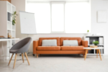 Blurred view of modern office with shelving units, workspace and cozy orange sofa near window