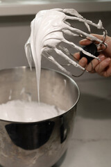 A pastry chef skillfully prepares whipped cream mixed with meringue in a shiny metal bowl, showcasing the smooth texture and glossy finish at a well-equipped cooking space.