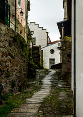 A narrow, wet cobblestone street winds between stone walls and buildings, leading towards a white building with a unique stepped design.