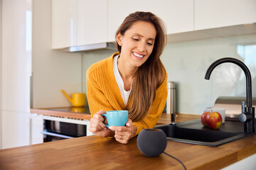 Woman in a kitchen talking to smart speaker while having coffee