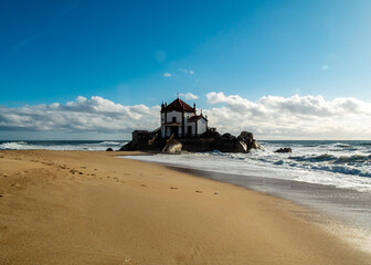 A chapel on a rocky outcrop on the beach with waves crashing against it.