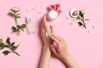 Female hands with jar of cosmetic cream, bottle of oil and beautiful rose flowers on pink background