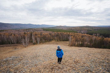 Person in blue walking toward forest, Individual with outdoor gear moves into dense woods, Adventurer dressed in functional clothing advances through grassy hillside toward forested