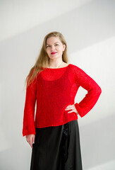 young blonde smiling beautiful lady in red handmade sweater and black skirt and black flower on the neck posing in white studio