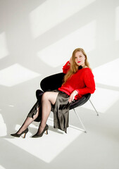 young blonde smiling beautiful lady in red handmade sweater and black skirt and black flower on the neck sitting on a chair