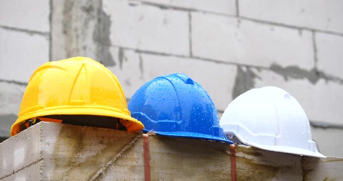 Raindrops on yellow, blue, white safety helmet placed on pile of bricks with house under construction background, construction problem concept.