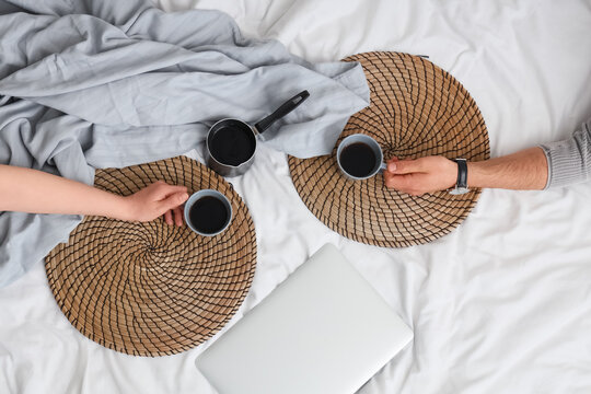 Young couple with cups of coffee, cezve and laptop on bed