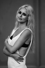 Young woman with long blonde hair, wearing a stylish white dress, poses confidently with a soft smile, showcasing elegance and beauty in a monochrome portrait
