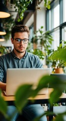 Young man working intently on laptop surrounded by plants in a modern cafe