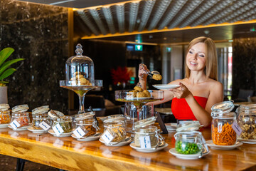 Elegant woman in red dress selecting cookies from dessert table in luxury hotel buffet setting with various snacks in jars. Hotel lounge