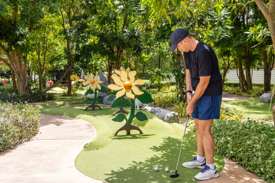 Man playing mini-golf in outdoor garden setting with large flower decorations, focusing on putting technique in sunny park. - Powered by Adobe