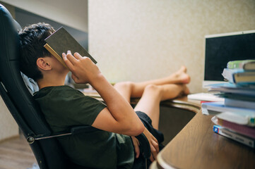 Teenager resting at desk with book on face feeling tired from studying