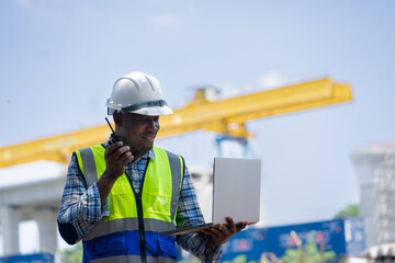 Construction Manager Using Radio and Laptop to Direct Site Work,African American Engineer Communicating Orders on a Job Site,Supervisor with Hard Hat and Walkie-Talkie looking Infrastructure Project.