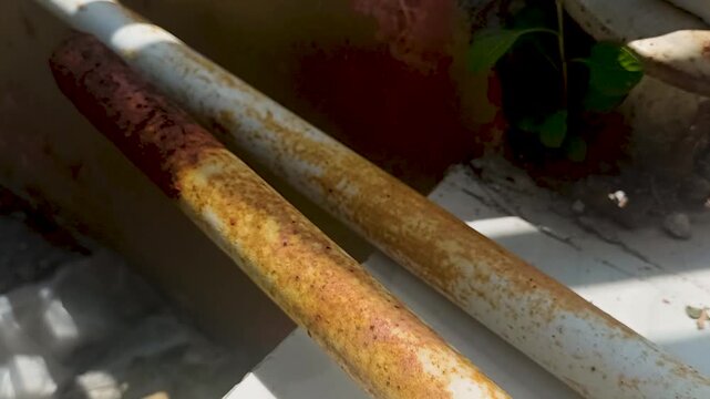 Close up view of two parallel metal pipes with peeling white paint and rust, casting shadows on a concrete surface, suggesting corrosion and weathering effects on urban infrastructure.