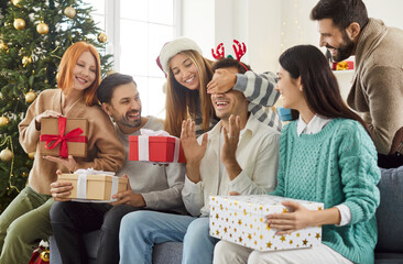 Group of happy young people closing eyes of their friend, making him surprise on Christmas. Smiling...