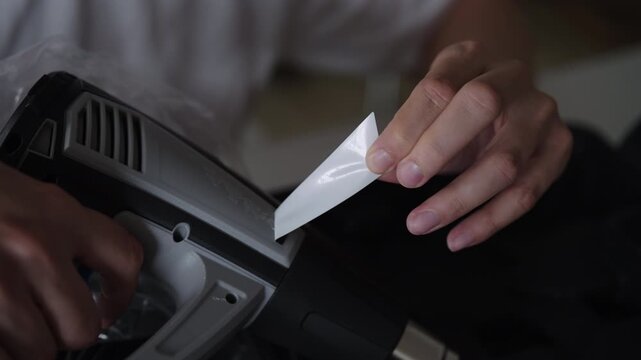 Close-up of male hands removing white label or sticker from dark gray electronic device hot air blower, showing process of sticker removal and product customization. Shooting in slow motion.