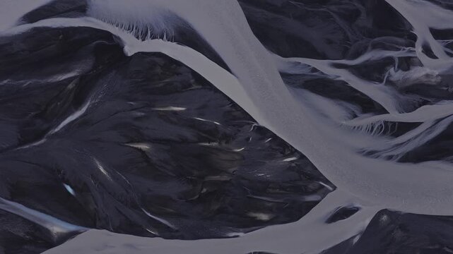 Drone pans and tilts above braided glacial rivers in Iceland as milky meltwater splits and rejoins over black sand and sediment under soft diffuse daylight.