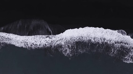 Top down aerial view shows black sand and white surf on an Icelandic shore near Vik. Waves roll and break as the camera drifts parallel, forming lace like foam patterns.