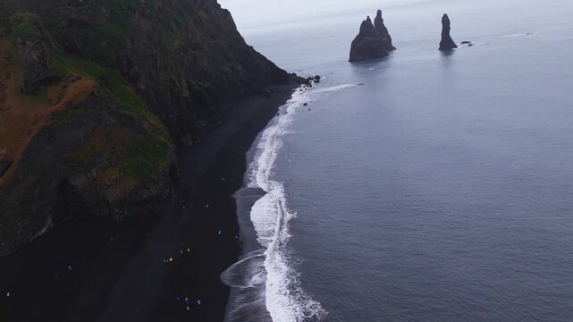 Aerial view pans along Reynisfjara near Vik, Iceland. Black sand, basalt cliffs, sea stacks, white surf, tiny figures, tents. Overcast light and slow lateral motion.