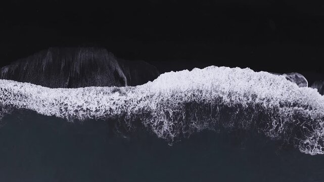 Top down aerial view shows black sand and white surf on an Icelandic shore near Vik. Waves roll and break as the camera drifts parallel, forming lace like foam patterns.