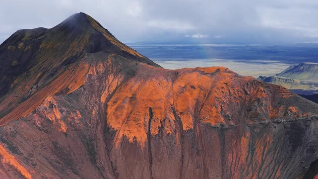 Aerial view moves across a steep cone and crater rim in Iceland highlands, near Landmannalaugar, with orange mineral streaks, deep gullies, layered strata, and a faint rainbow.