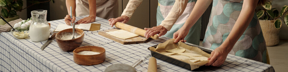 Senior Caucasian woman rolling dough with young adult Caucasian women preparing pastry together on kitchen table, hands visible working with baking ingredients and utensils