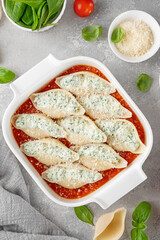 Raw conchiglioni pasta stuffed ricotta cheese and spinach with tomato sauce and parmesan cheese on top in a white baking dish on a gray concrete background.
